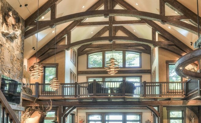 Interior of a Custom Timber Frame Residence in WI with dark stained, arched king post trusses, cathedral windows and ceilings, and a stone fireplace.