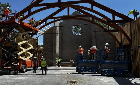 Local construction crews install a heavy timber truss being lowered into place by a crane.