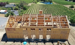 Wood-framed two-story house under construction in a rural farm setting, with scaffolding and exposed framing; vineyards in the background.
