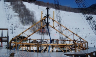 Wooden triangular frame under construction on a snow-covered slope, with cranes in the background.