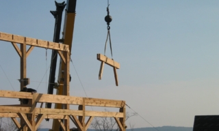 Crane lifts a wooden beam over a timber-framed construction site against a blue sky.